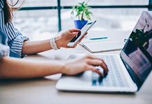 Person using a laptop while holding a smartphone at a desk.