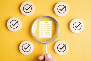 Hand holding a magnifying glass over a checklist, surrounded by circular checkmark icons on a yellow background, symbolizing review or verification.