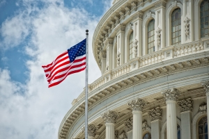 The dome of the United States Capitol building against a blue sky with clouds, with an American flag waving in the foreground, representing the U.S. government or Congress.
